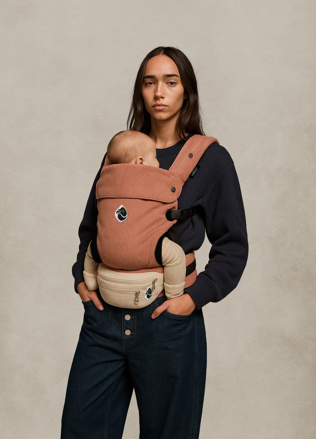 Model trägt die Velia Nuvola Cord Babytrage in Rosé Muse mit Essenza Bag in Weiß – feminine Eleganz und ergonomischer Komfort in einem High-Fashion-Look.